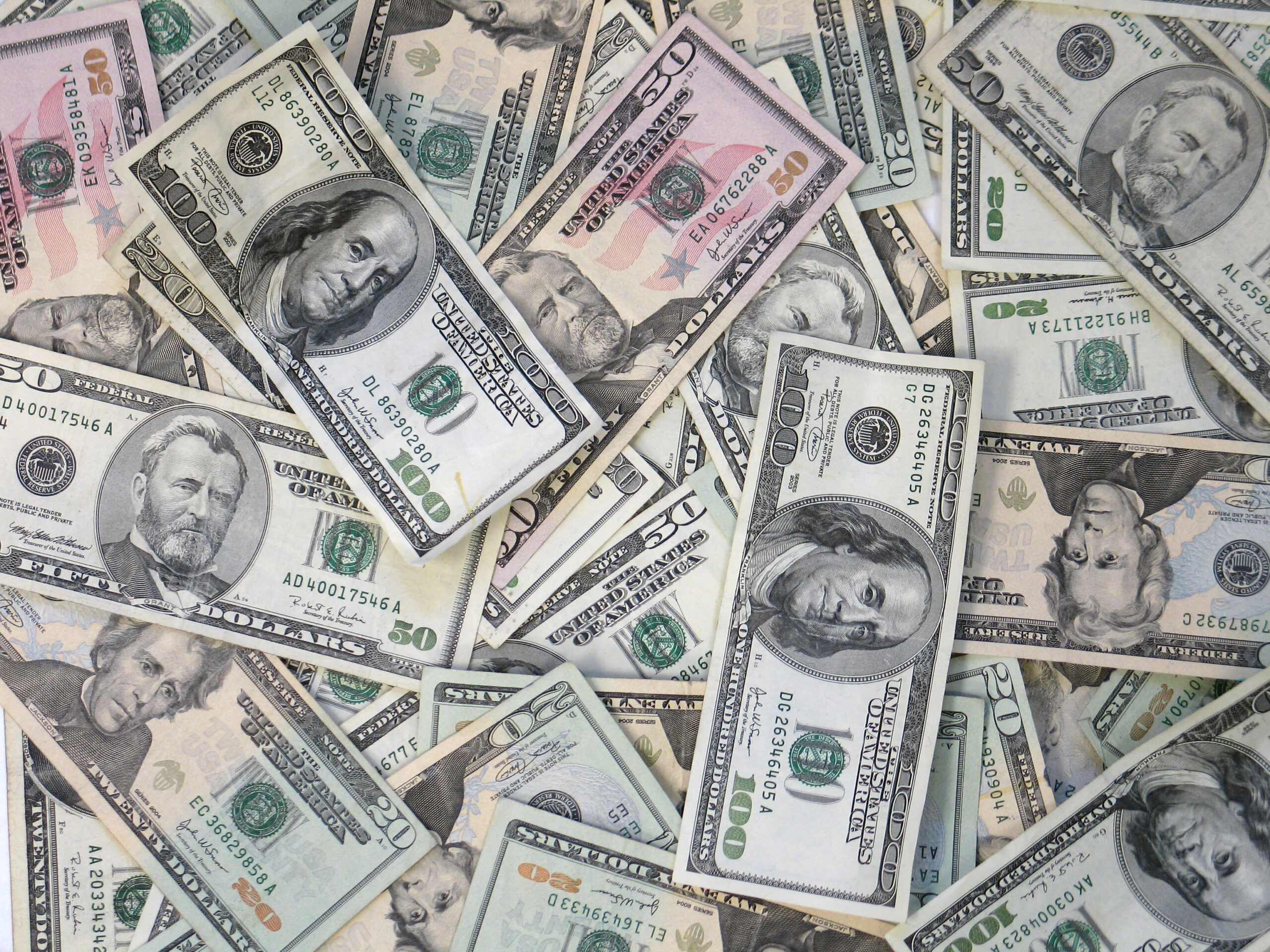 A scattered pile of U.S. dollar bills of various denominations, including $100, $50, $20, $10, and $1 notes, showing different portraits and designs.