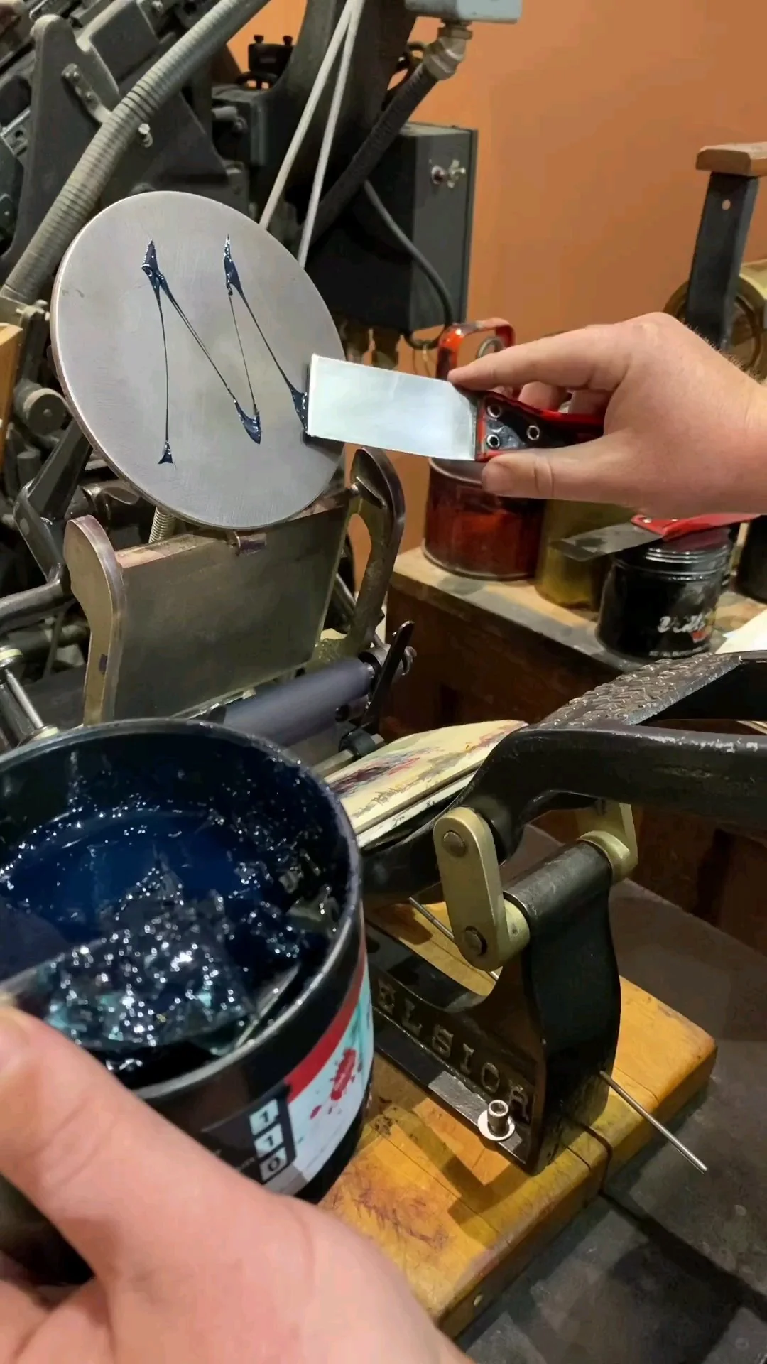 A person spreads dark blue ink onto the round plate of a vintage letterpress machine using a spatula. An open ink can and various printing supplies are visible in the background.