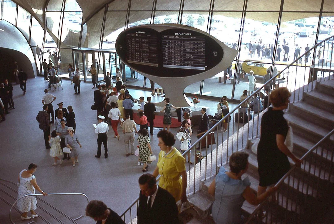 Crowd inside a mid-20th-century airport terminal with a large split-flap departure board, people walking, and others descending a curved staircase. Sunlight streams in through large windows.