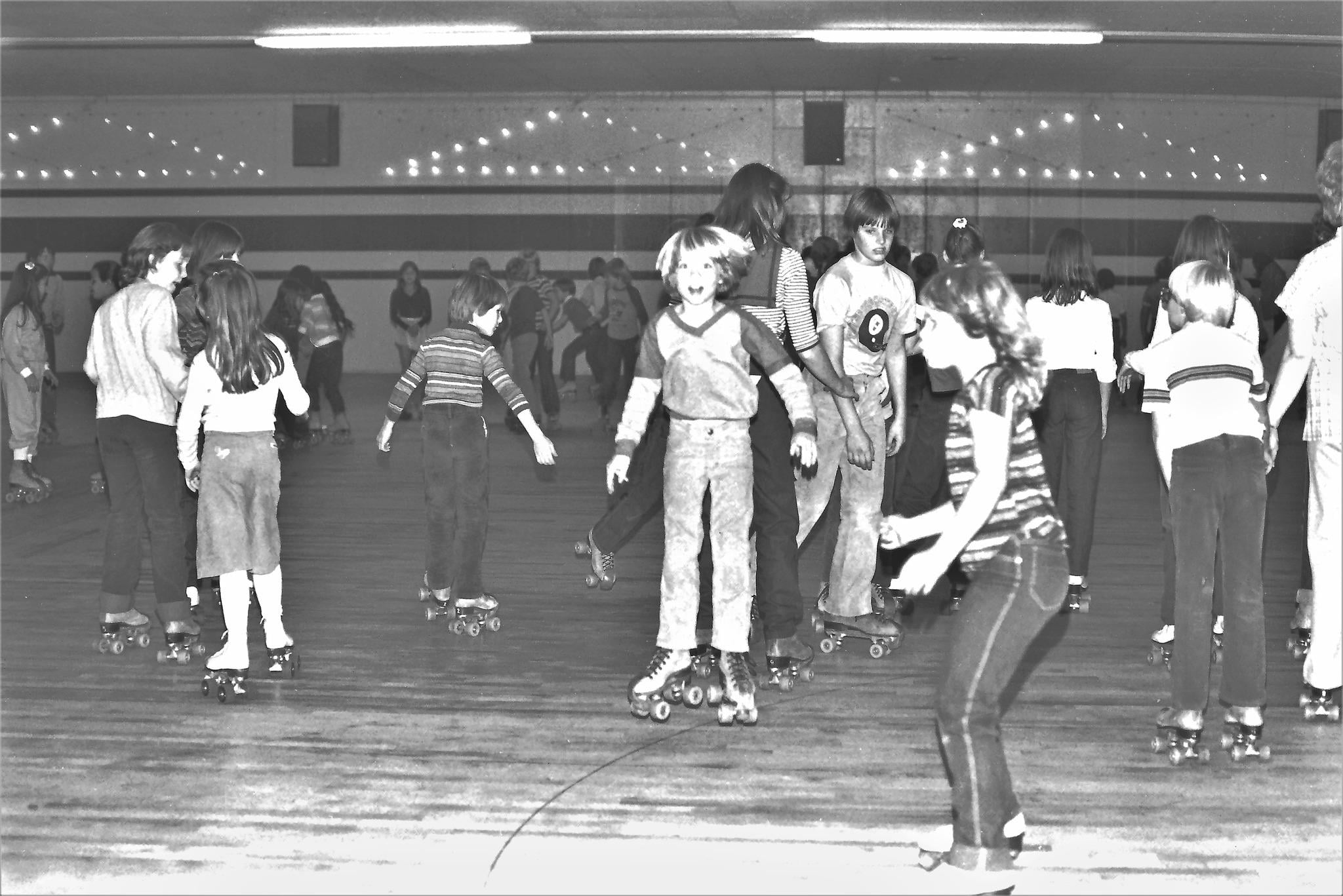Children roller skating indoors at a rink, some holding hands, others skating alone. The scene is lively with kids of various ages, and adults are visible in the background watching.