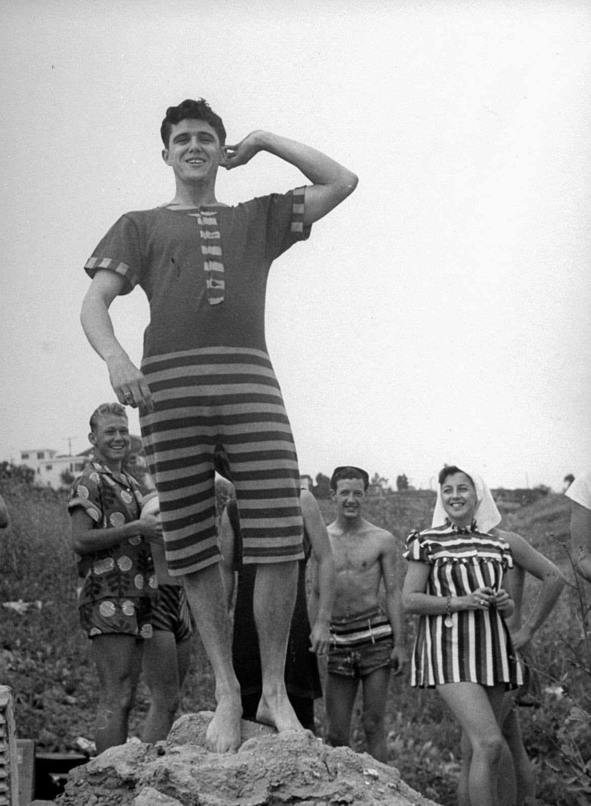 A smiling man poses on a rock in a vintage striped swimsuit, flexing his arm, while several people in old-fashioned swimwear stand and smile in the background outdoors.