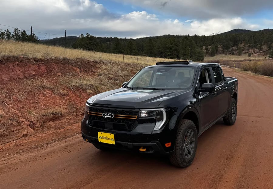 A black Ford pickup truck is parked on a red dirt road beside grassy terrain, with trees and hills visible in the background under a partly cloudy sky.