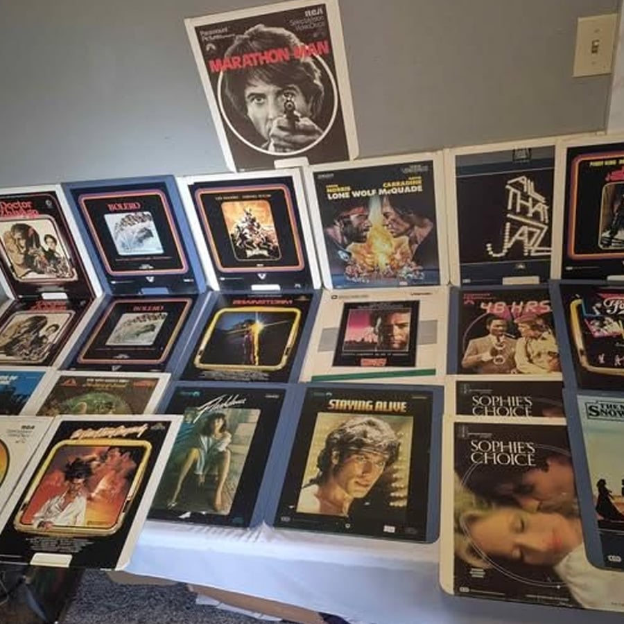 A table displays vintage movie laserdiscs in their sleeves, featuring various classic film covers. A poster for “Marathon Man” is on the wall above the collection.