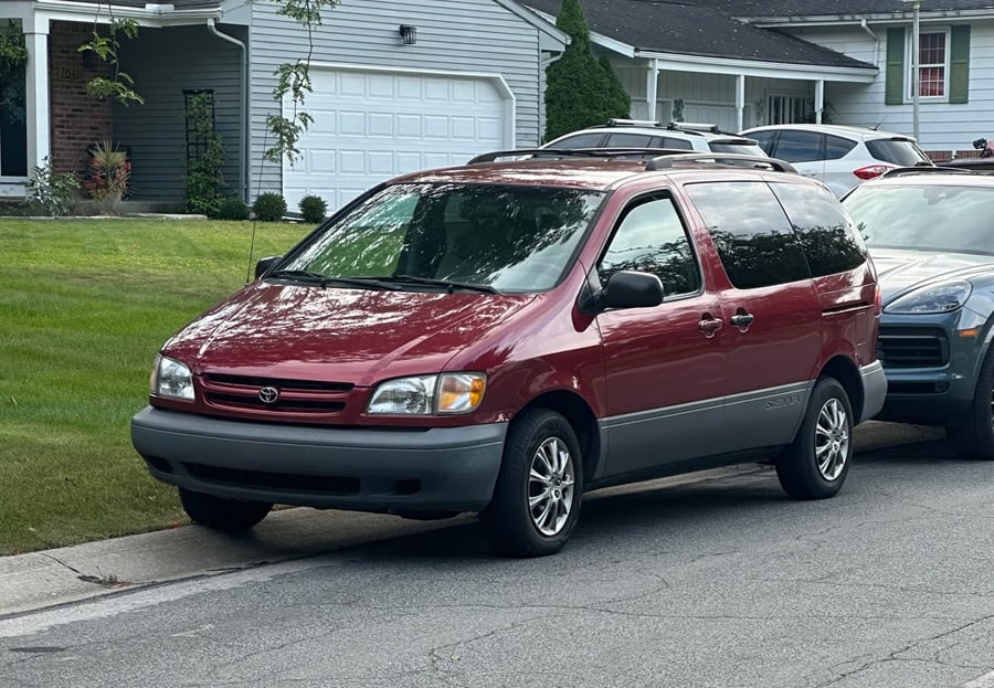 A red Toyota Sienna minivan is parked on a suburban street in front of a gray house with white trim and a white garage door. Another car is parked closely behind it. The surroundings are green and residential.