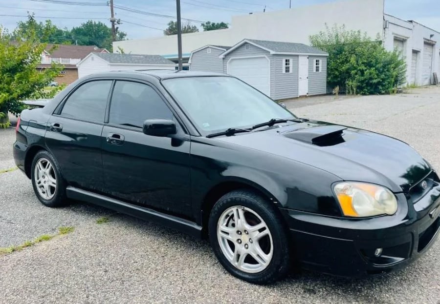 A black sedan with tinted windows and alloy wheels is parked on a gravel lot near a small gray shed and some trees.
