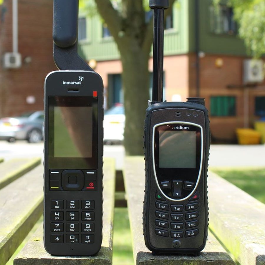 Two rugged satellite phones with antennas extended are standing upright on a wooden bench outside, with buildings and trees blurred in the background.