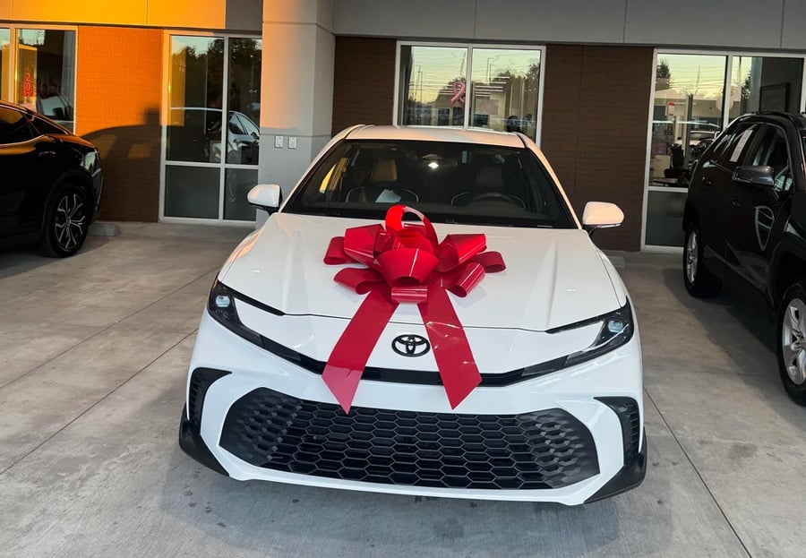 A white Toyota sedan with a large red bow on its hood is parked outside a dealership, ready for delivery.