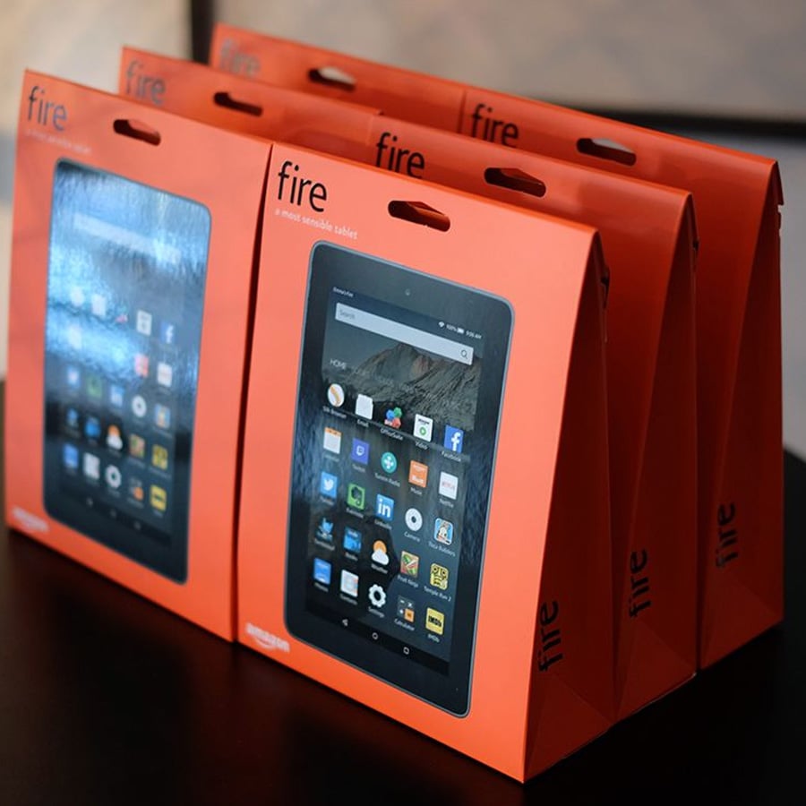 Four Amazon Fire tablets in orange packaging are displayed upright on a black surface. The front of each box shows the tablet screen with various app icons visible.