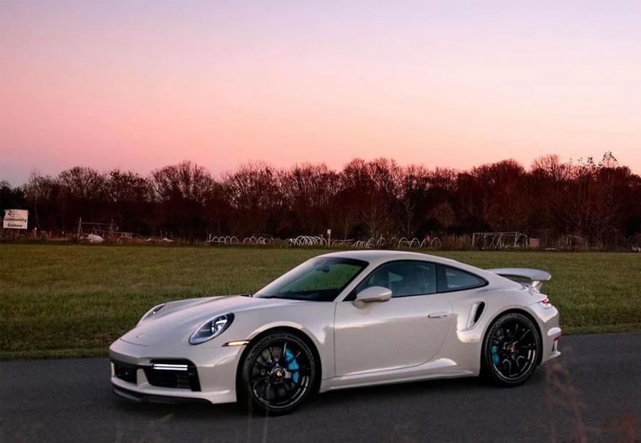 A white sports car parked on a road near a field at sunset, with trees and a pink-purple sky in the background.