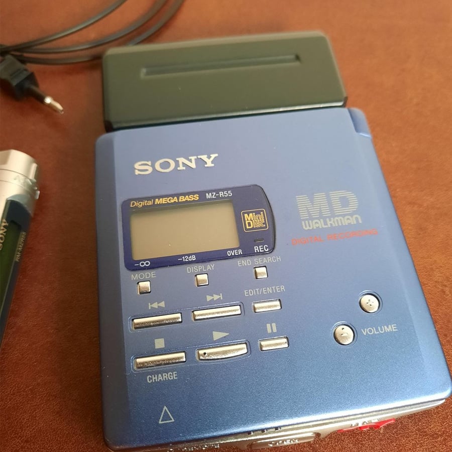 A blue Sony MD Walkman digital recorder with various buttons and a display screen is shown on a brown surface, with a black headphone cable and battery partly visible beside it.