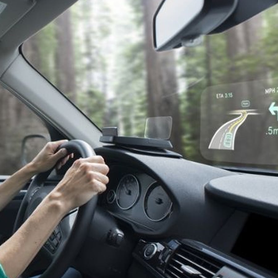 Person driving a car through a forested area with a digital navigation heads-up display projected onto the windshield showing route, ETA, and speed.