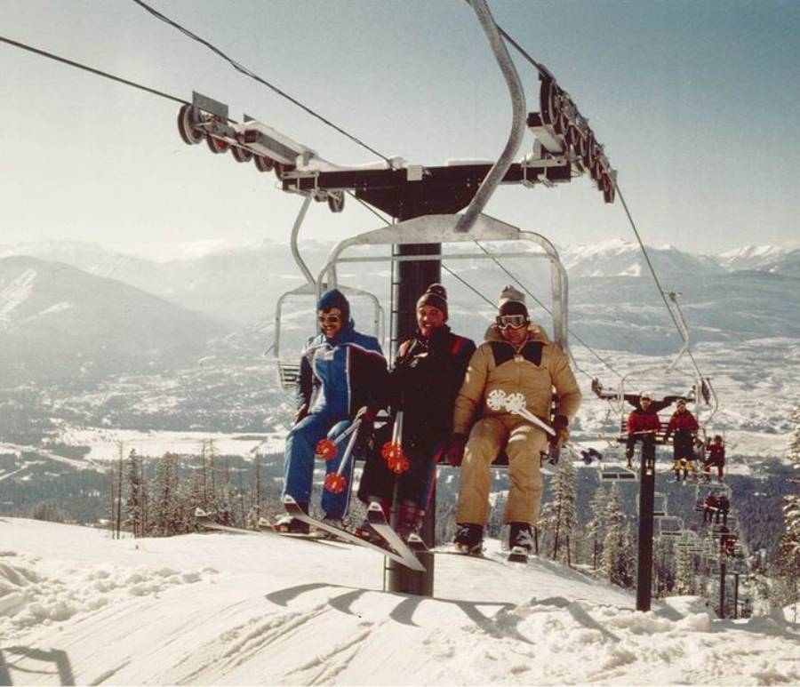 Three people in colorful ski suits ride a chairlift over a snowy mountain slope, with ski poles in hand. Snow-covered peaks and a valley are visible in the background under a clear sky.