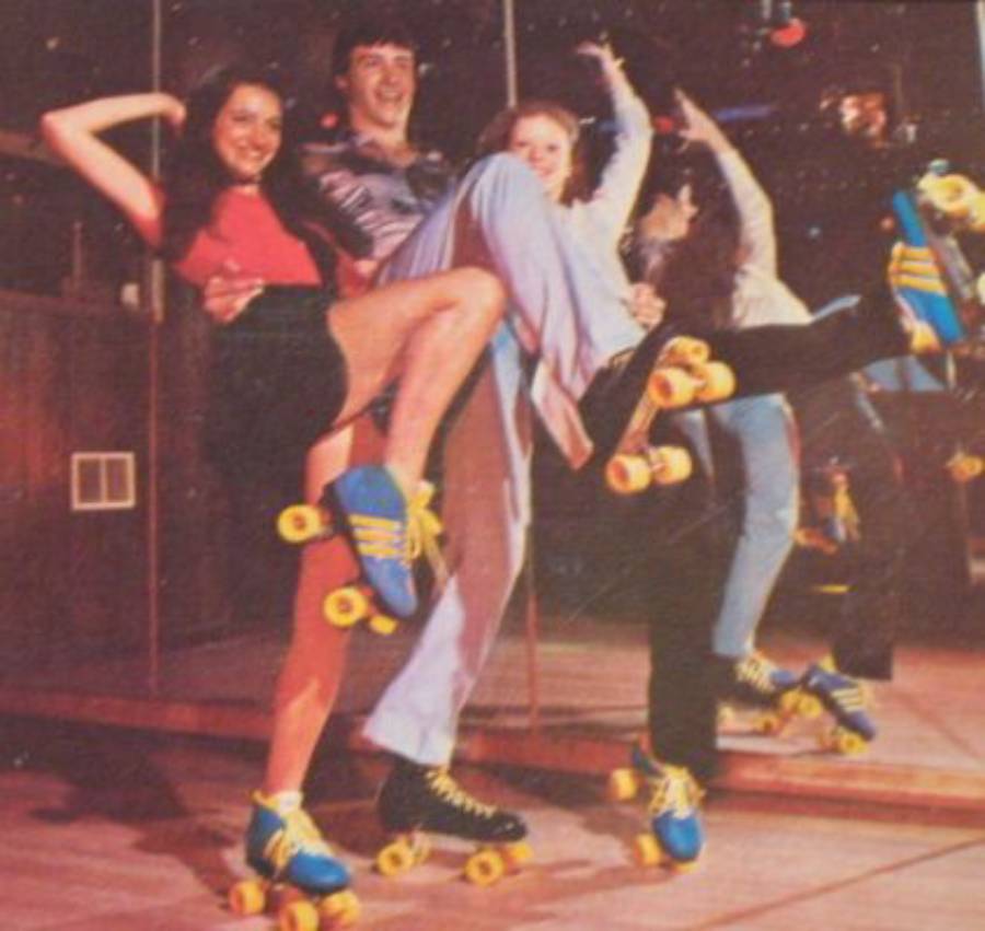 Three people in retro outfits pose playfully on roller skates in front of a mirror, lifting their legs and smiling. Bright yellow and blue skates are visible on all three.