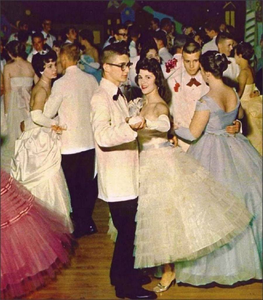 Teenagers in formal attire dance at a prom. The girls wear colorful, full-skirted dresses, and the boys wear white jackets with bow ties. The setting is festive, with couples smiling and enjoying the evening.