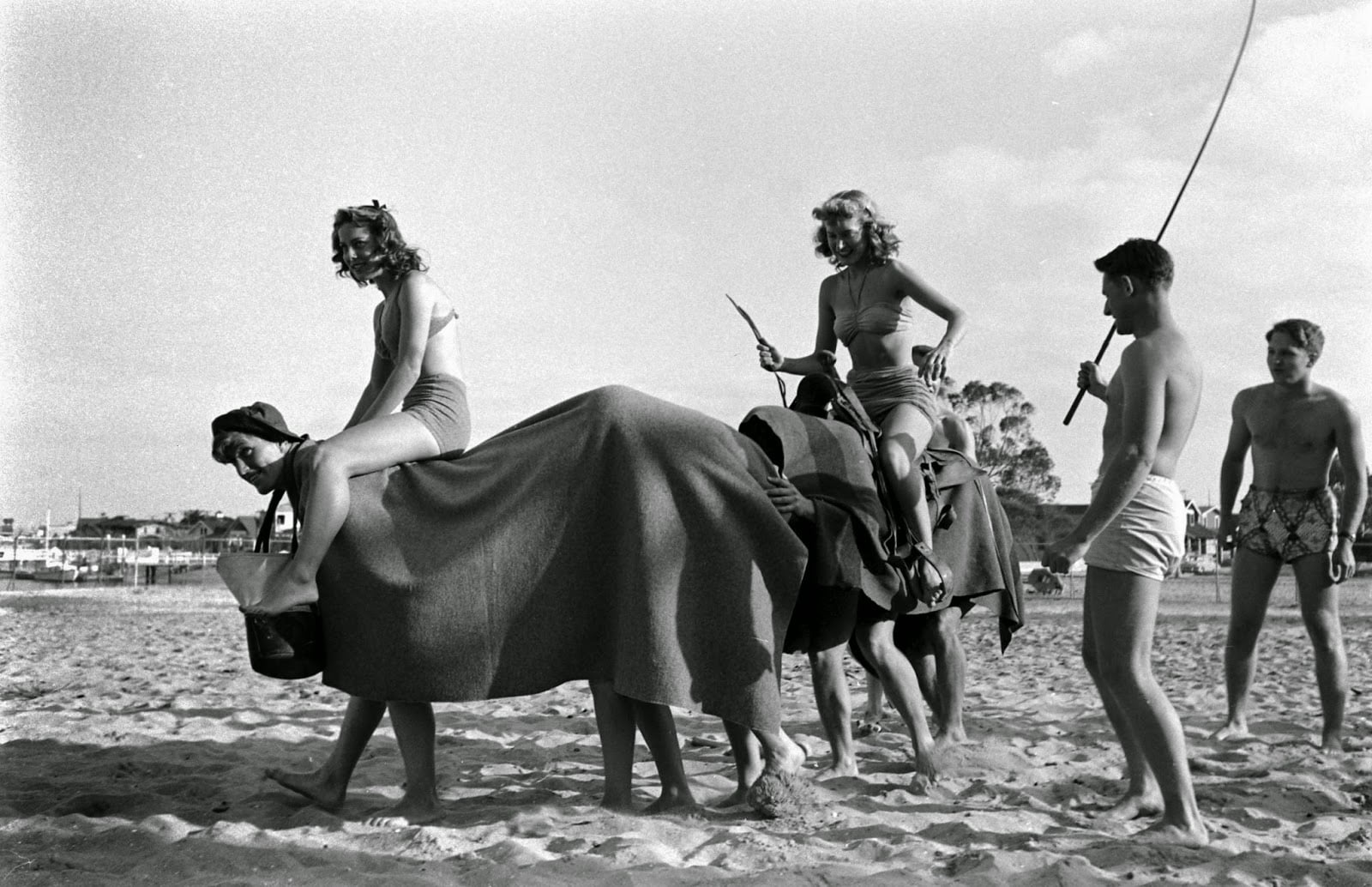 Two women in swimsuits ride a “camel” made of two people covered by a blanket, while two men in swimsuits walk beside them on a sandy beach. One woman pretends to hold reins and a stick.