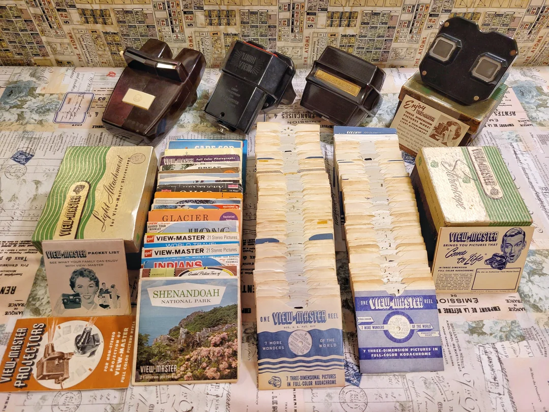 Four vintage View-Master viewers and stacks of View-Master reels with original boxes are displayed on a decorated table, featuring travel and scenic reel themes.