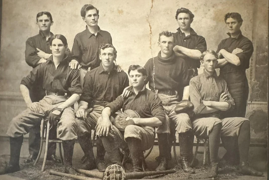 A vintage black-and-white photo of nine young men in athletic uniforms, posing in two rows. Baseball bats and a glove rest on the floor in front of them.