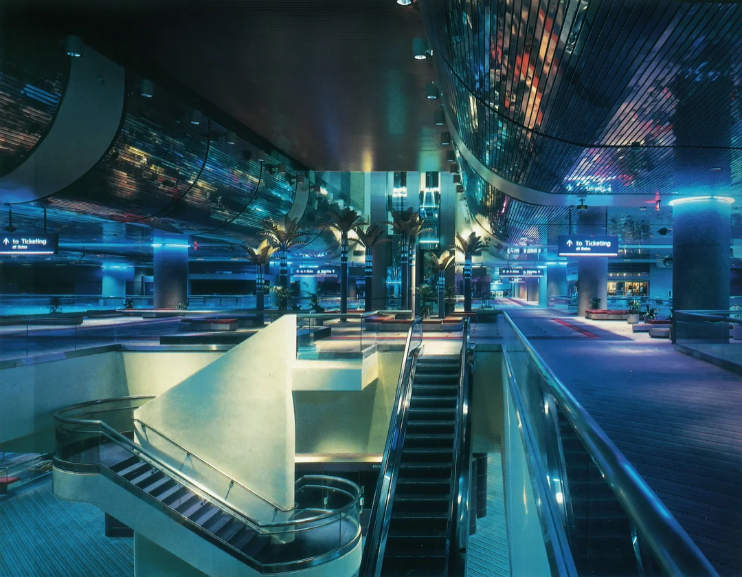 A modern, futuristic airport terminal with blue lighting, escalators, palm trees, and reflective surfaces. Signs for ticketing and baggage claim hang from the ceiling, creating a sleek, high-tech atmosphere.