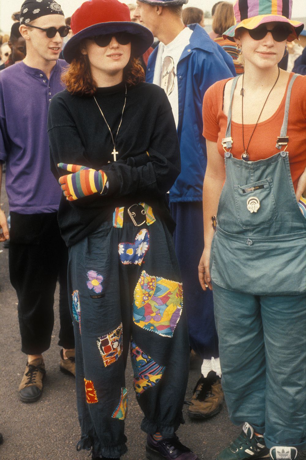 A group of young people in colorful, eclectic 1990s clothing, including hats, patchwork pants, and striped gloves, stand together outdoors at what appears to be a festival or event.
