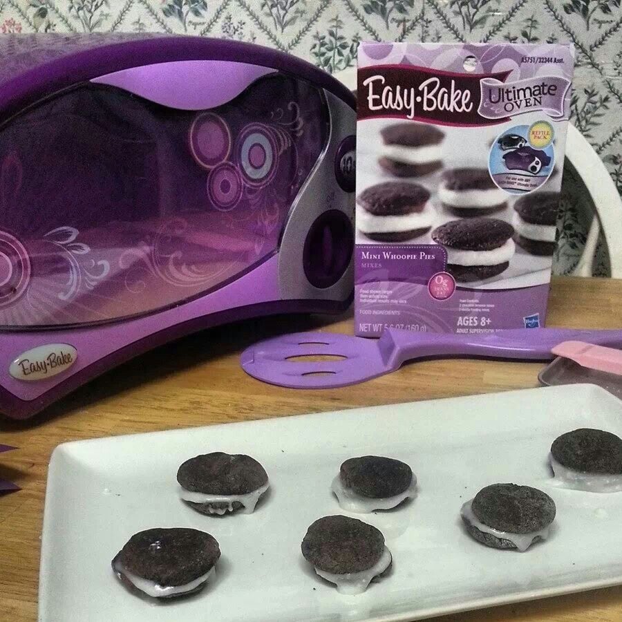 A purple Easy-Bake Oven, a spatula, a whoopie pie mix box, and six small whoopie pies with white filling on a plate are displayed on a table.