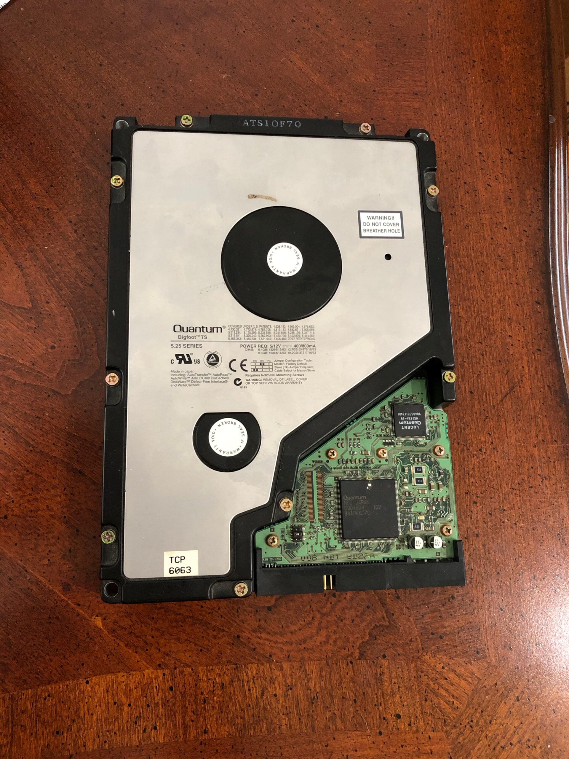 A Quantum hard drive with exposed circuit board sits on a wooden surface. Visible labels, screws, and electronic components are seen on the drive’s green PCB.