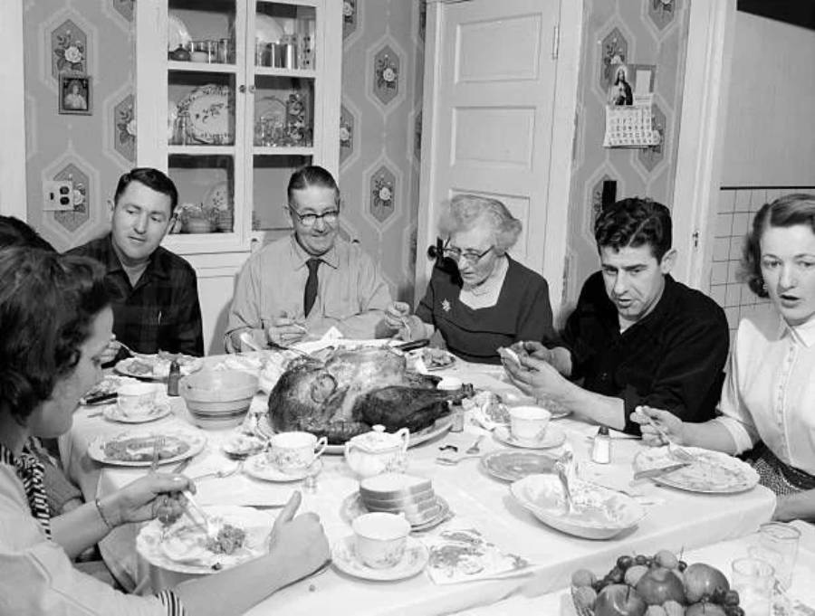 A family of six sits around a dining table, sharing a meal. There is a roasted turkey in the center, teacups, and plates of food. The setting appears to be a cozy, mid-20th century home.