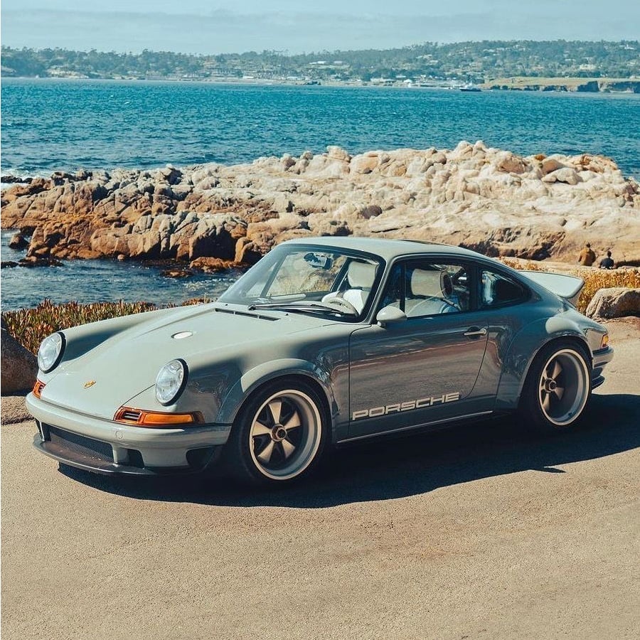 A classic silver Porsche sports car is parked by the ocean, with rocky shores and blue water in the background on a sunny day.