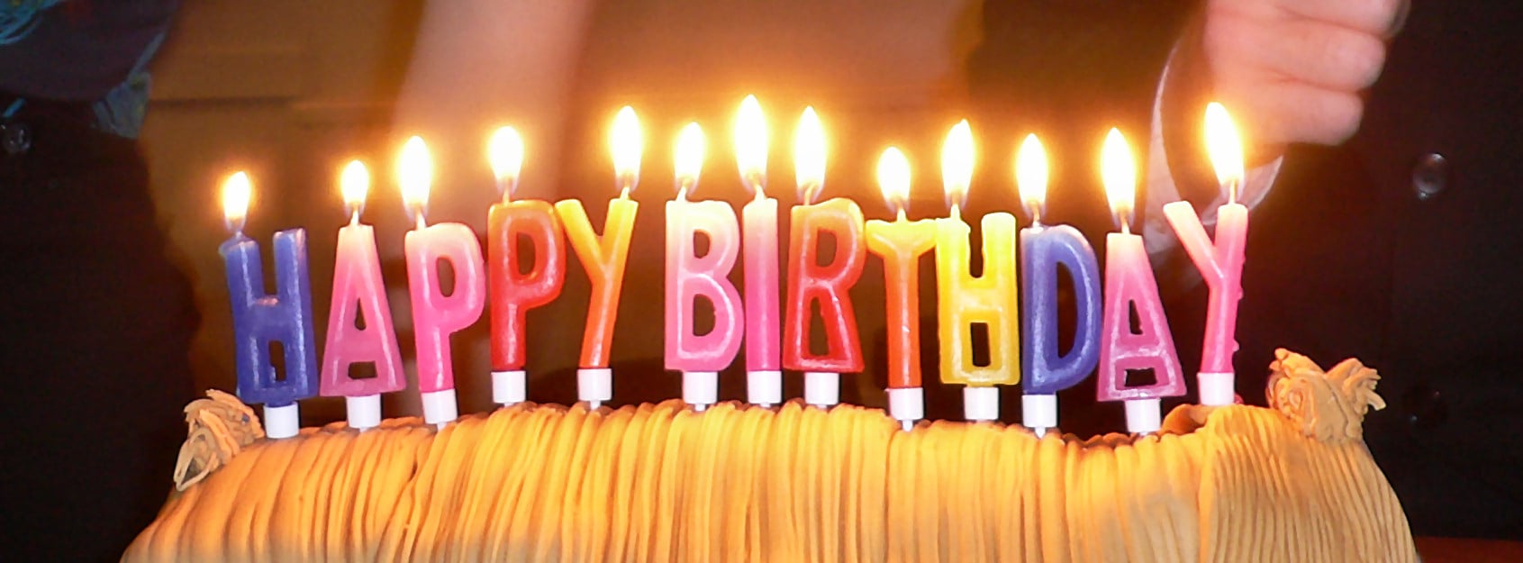 A birthday cake topped with lit candles shaped as colorful letters spelling “HAPPY BIRTHDAY.”
