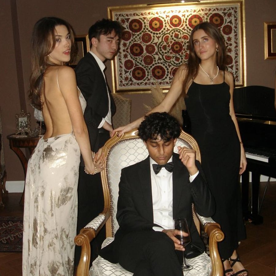 Four young adults in formal evening wear pose indoors; one man sits in an ornate chair holding a glass, while the other three stand behind him, two women in gowns and a man in a suit, all looking in different directions.