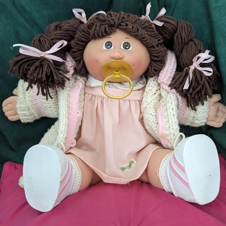 A soft doll with brown yarn hair in pigtails, pink bows, and a pink dress sits against a green background. The doll has a yellow pacifier, white shoes, and wears a knitted cream sweater.