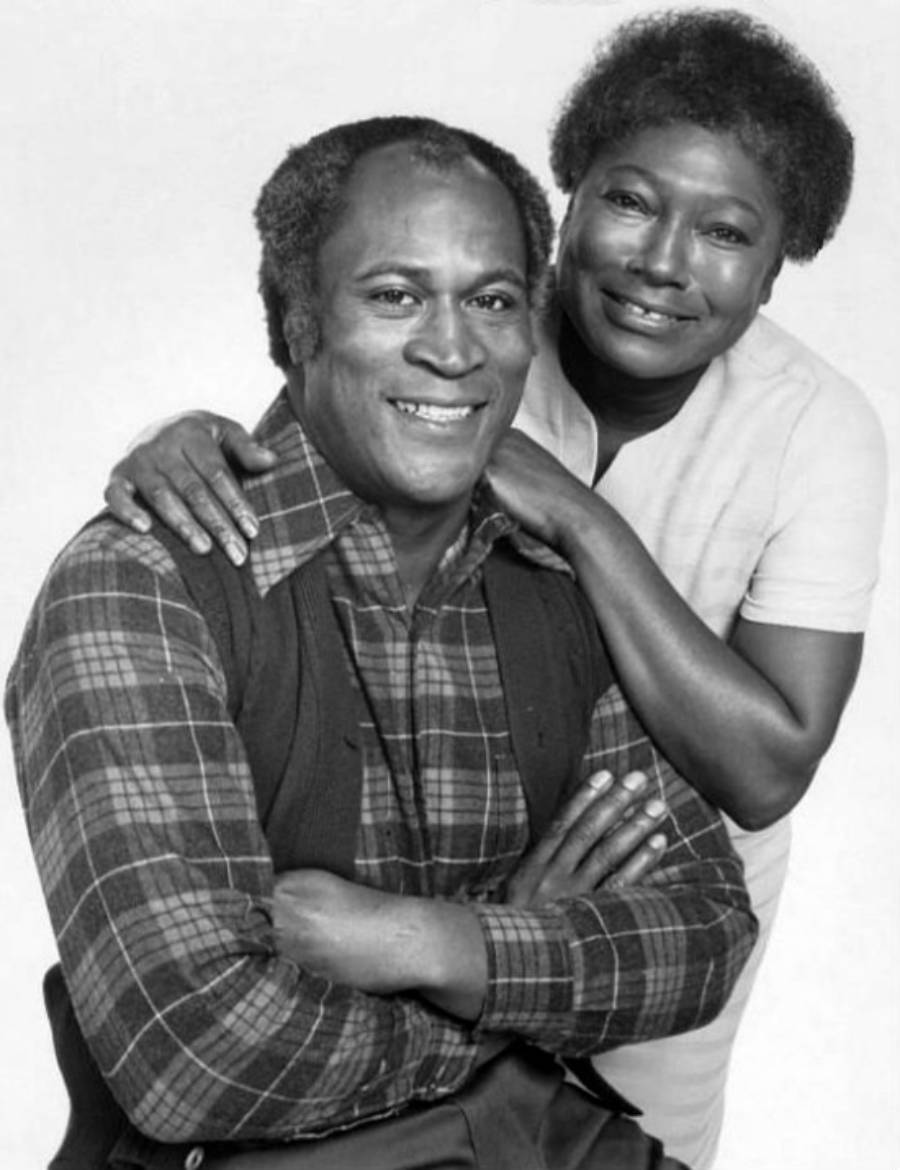 A smiling man in a plaid shirt and vest sits with his arms crossed as a woman in a light dress stands behind him, resting her hands on his shoulders. Both look at the camera against a plain background.