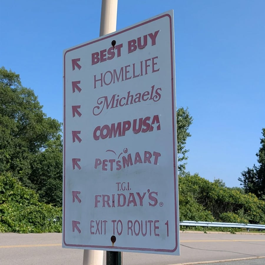 A sign lists businesses—Best Buy, Homelife, Michaels, CompUSA, PetSmart, T.G.I. Friday’s—with arrows pointing left and “Exit to Route 1” pointing right. Trees and blue sky are in the background.