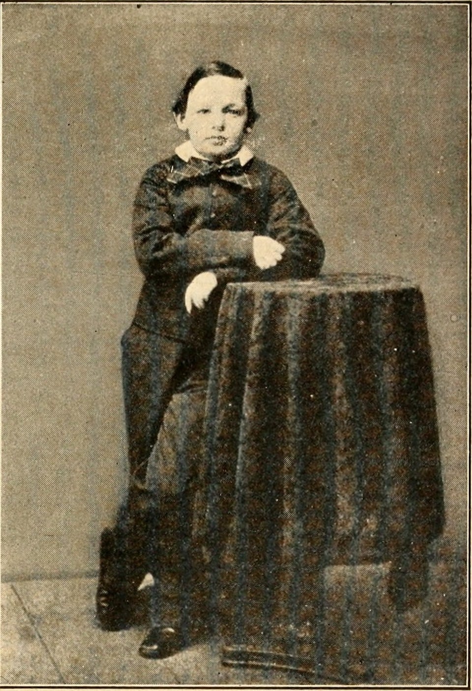 A young boy in formal clothing stands next to a draped table, resting one arm on it, in a vintage black and white photograph.
