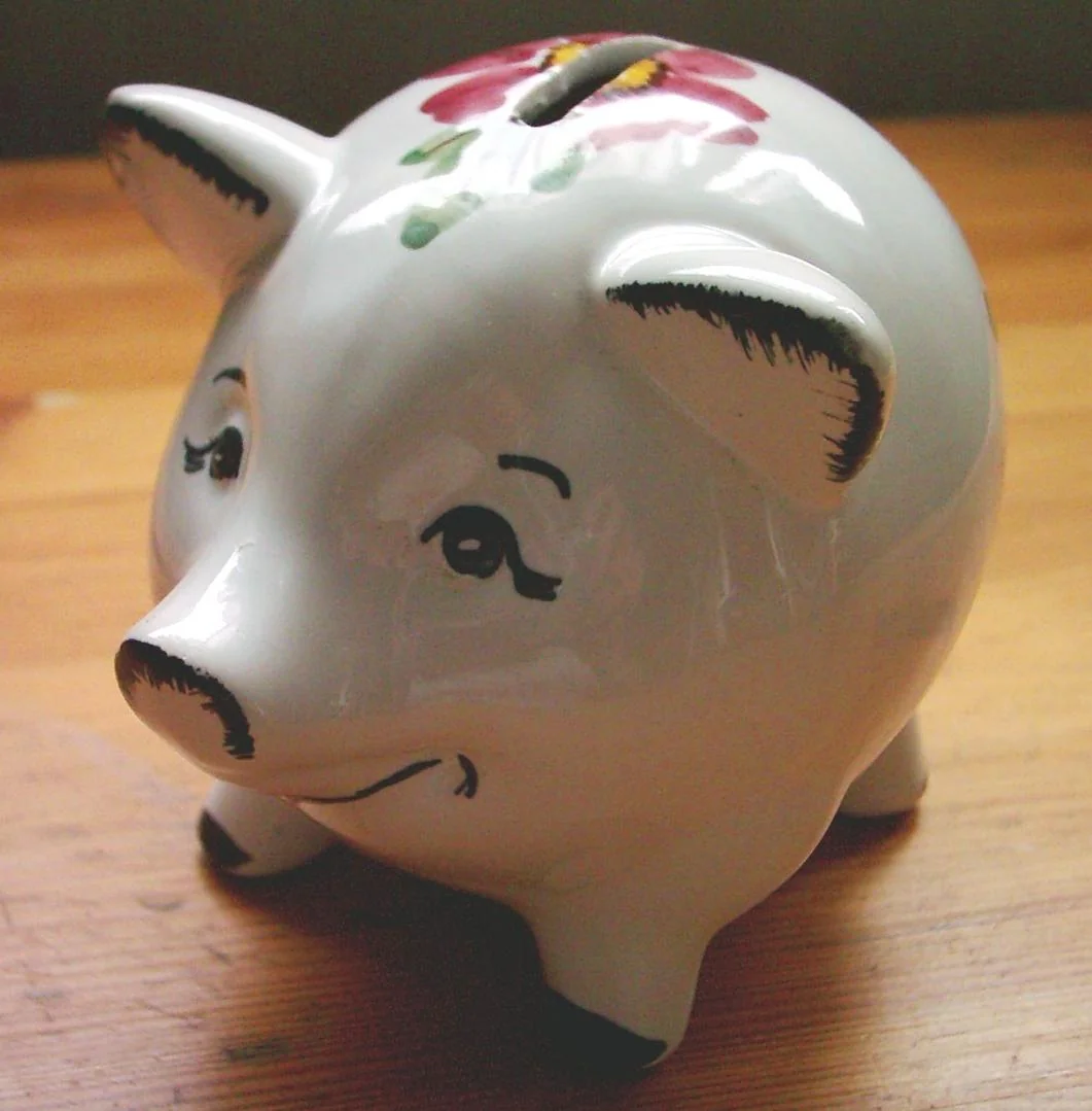A white ceramic piggy bank with a painted red flower design on its back sits on a wooden surface, facing slightly left.