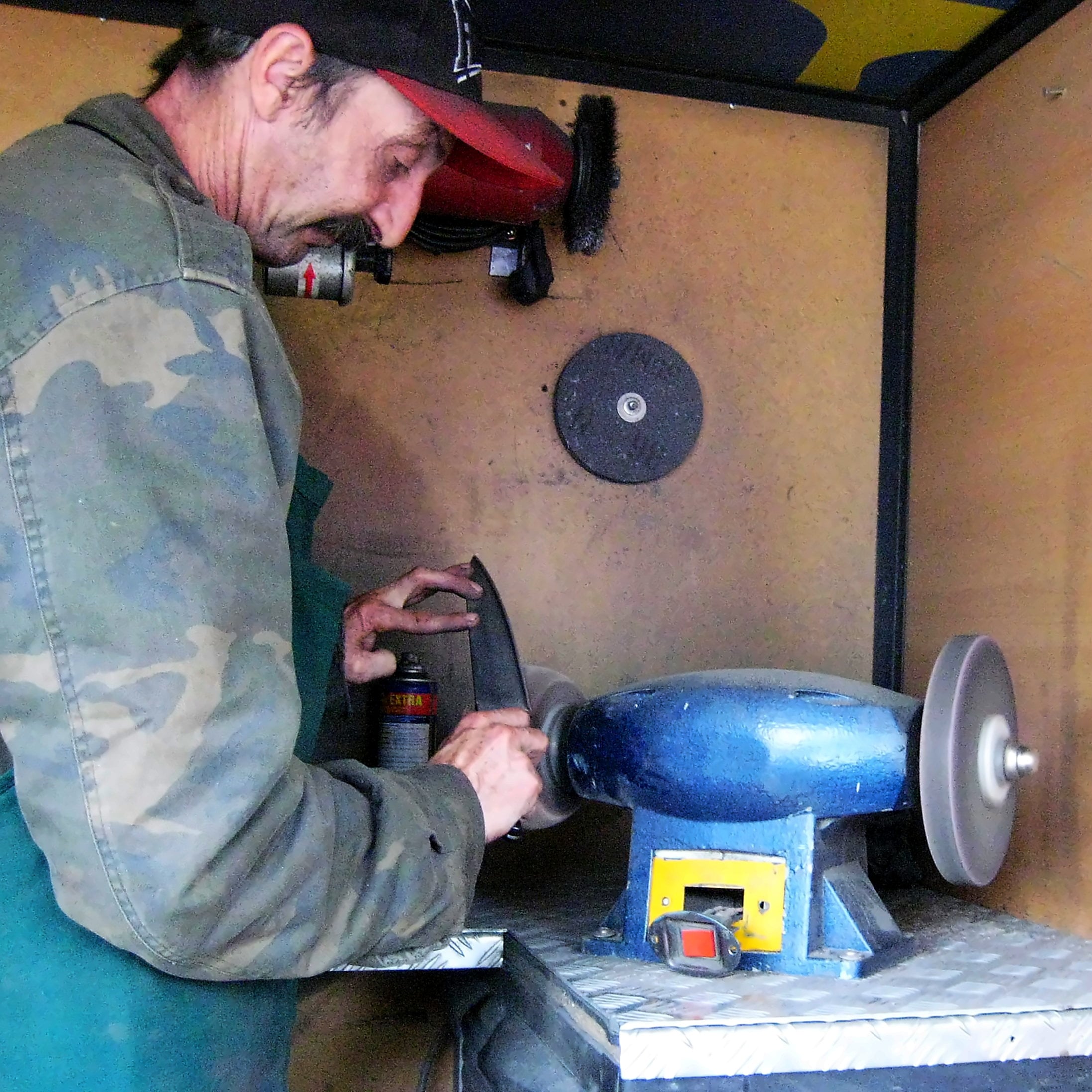 A man wearing a camouflage jacket uses a bench grinder to sharpen an iron in a small workshop. Tools and equipment are visible on the walls behind him.