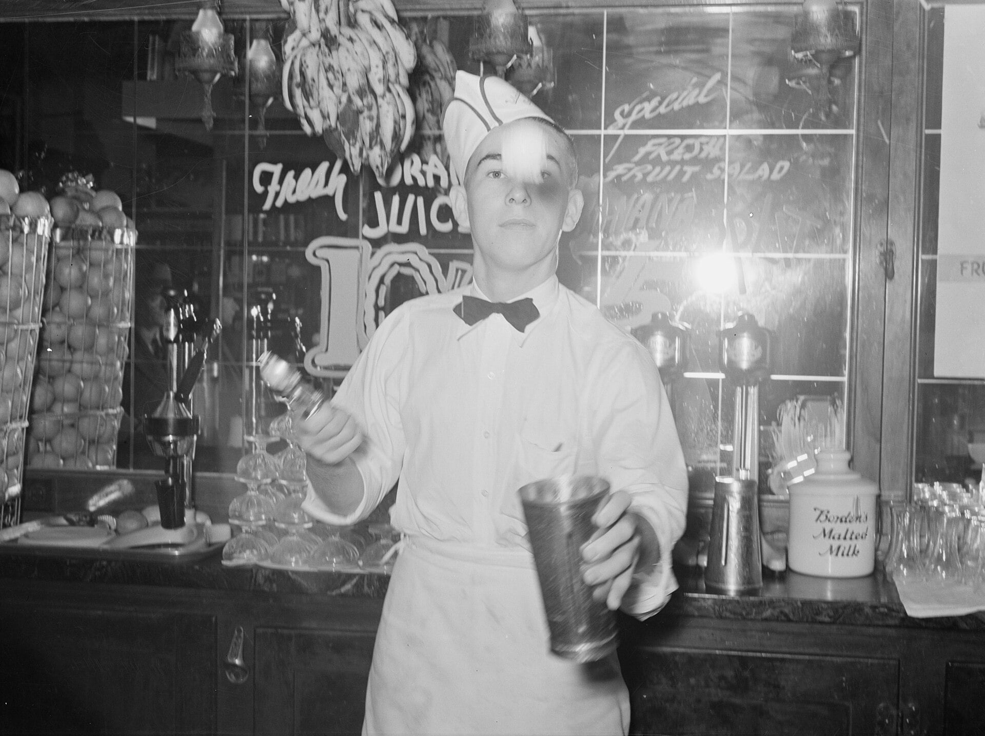 A soda jerk in a white uniform and hat prepares a drink behind a counter, surrounded by fruit, glassware, and milkshake equipment in a vintage diner or soda shop.
