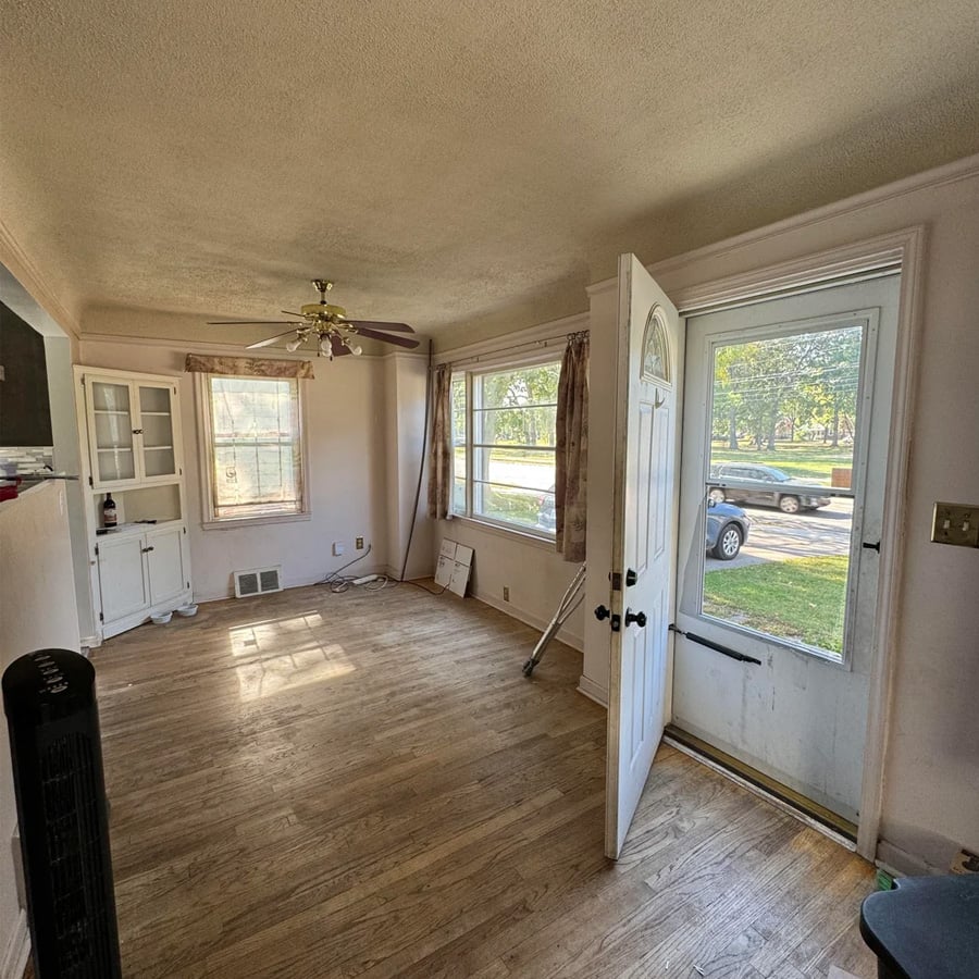 Bright, empty living room with wood floors, large windows, an open front door, ceiling fan, white cabinet, and sunlight streaming in. A parked car is visible through the windows.