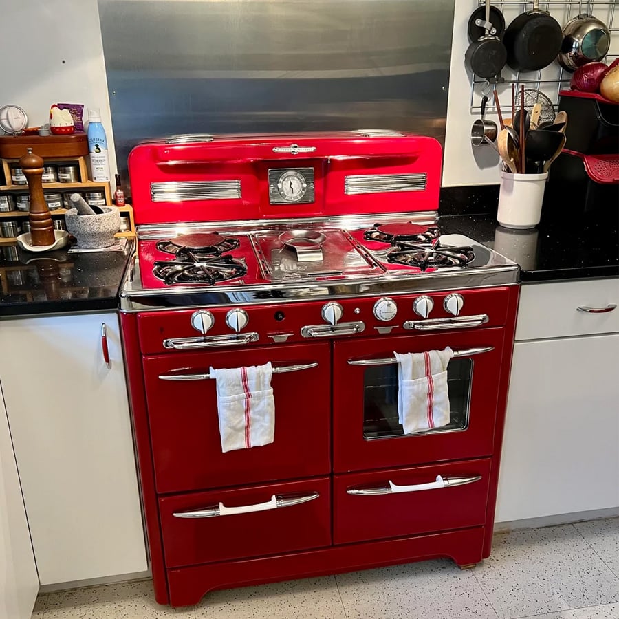 A vintage-style red stove with chrome accents, four burners, a central clock, two oven doors, and two striped dish towels hanging on the handles in a modern kitchen.