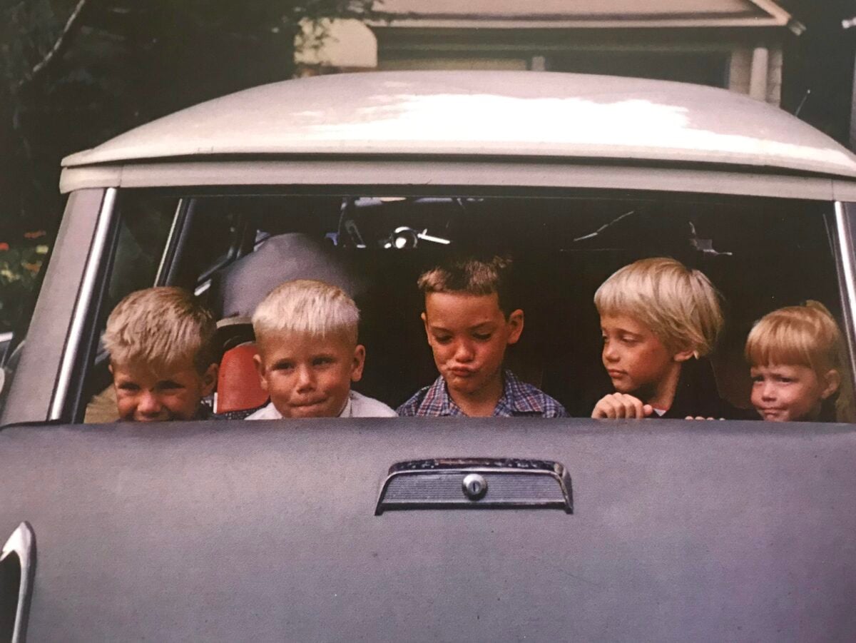 Five young children sit in the front seat of a vintage car, looking out the open window. The children have various expressions, and a house is visible in the background.