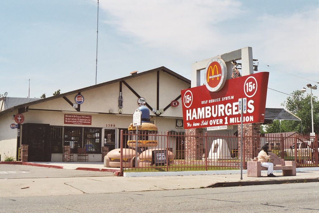 A vintage McDonald’s restaurant with a retro sign reading “Hamburgers 15¢” and “Over 1 million sold.” A person sits on a bench outside, and the building has a classic design with a beige exterior.