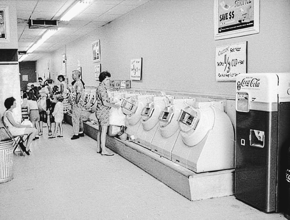 Black and white photo of people using washing machines at a laundromat. A Coca-Cola vending machine is on the right, with posters on the wall. Several adults and children stand or sit, waiting.