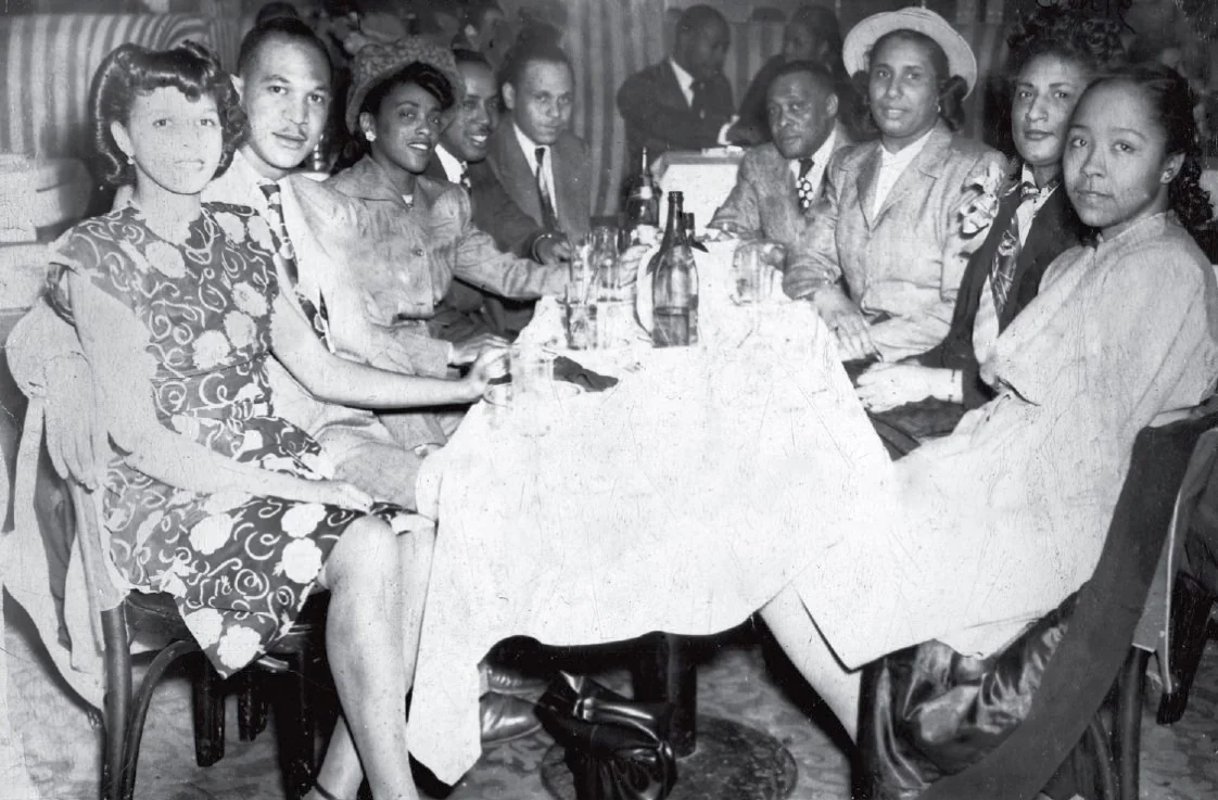 A group of men and women dressed in 1940s attire sit around a table with drinks at a lively social gathering in a restaurant or club.