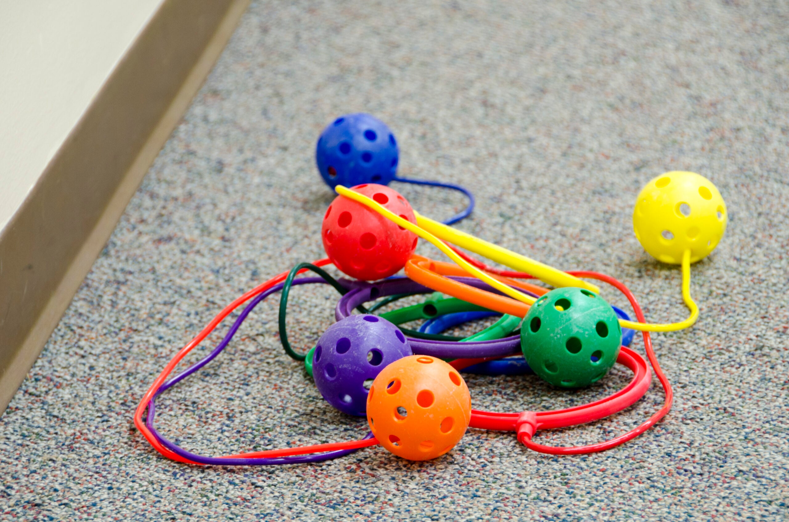 Colorful plastic balls with holes are attached to matching colored strings, lying on a speckled carpet near a beige wall and baseboard.