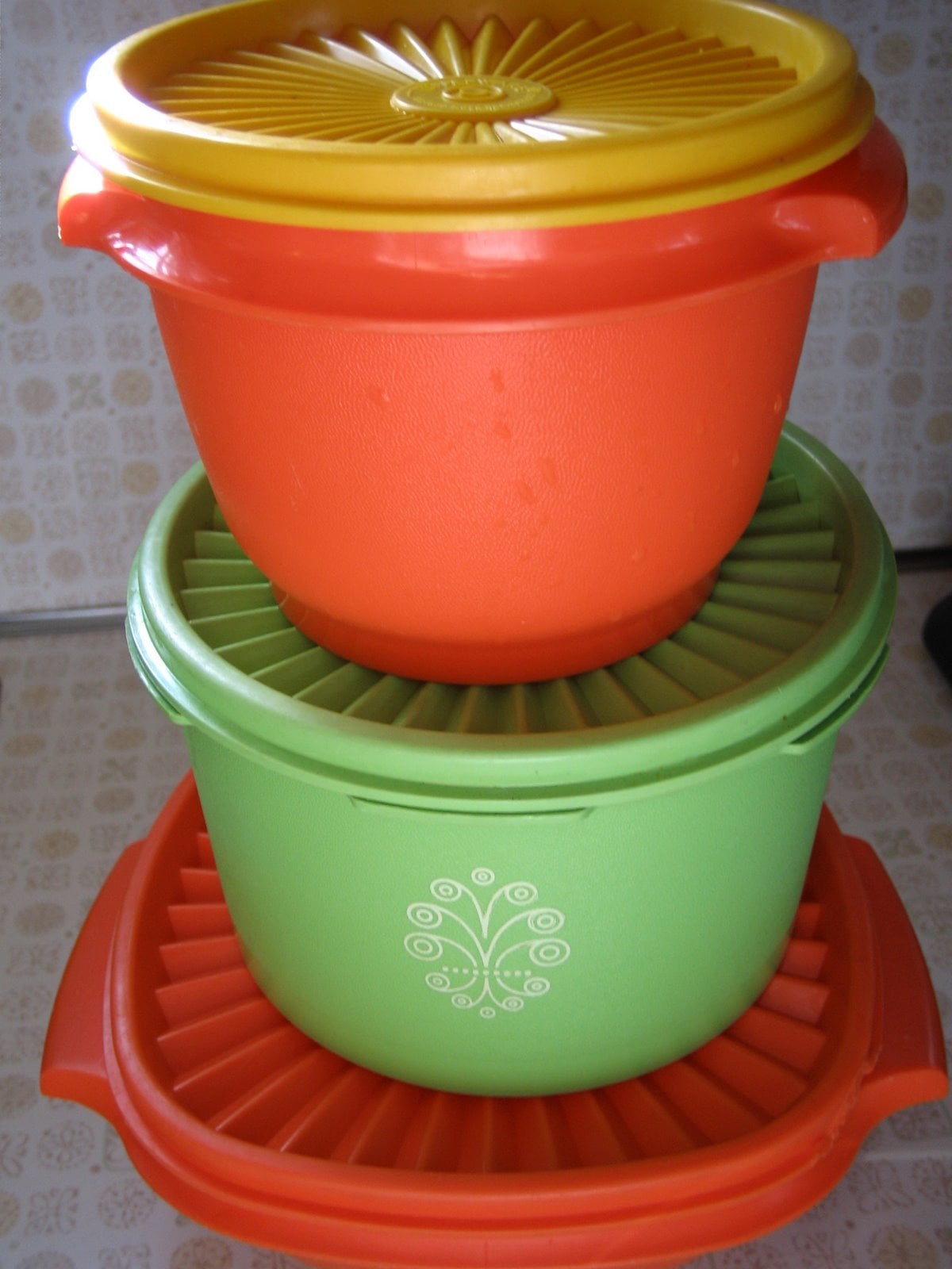 Three plastic containers are stacked; the top is orange with a yellow lid, the middle is green with a decorative white pattern and a green lid, and the bottom is orange with a matching orange lid, all on a patterned surface.