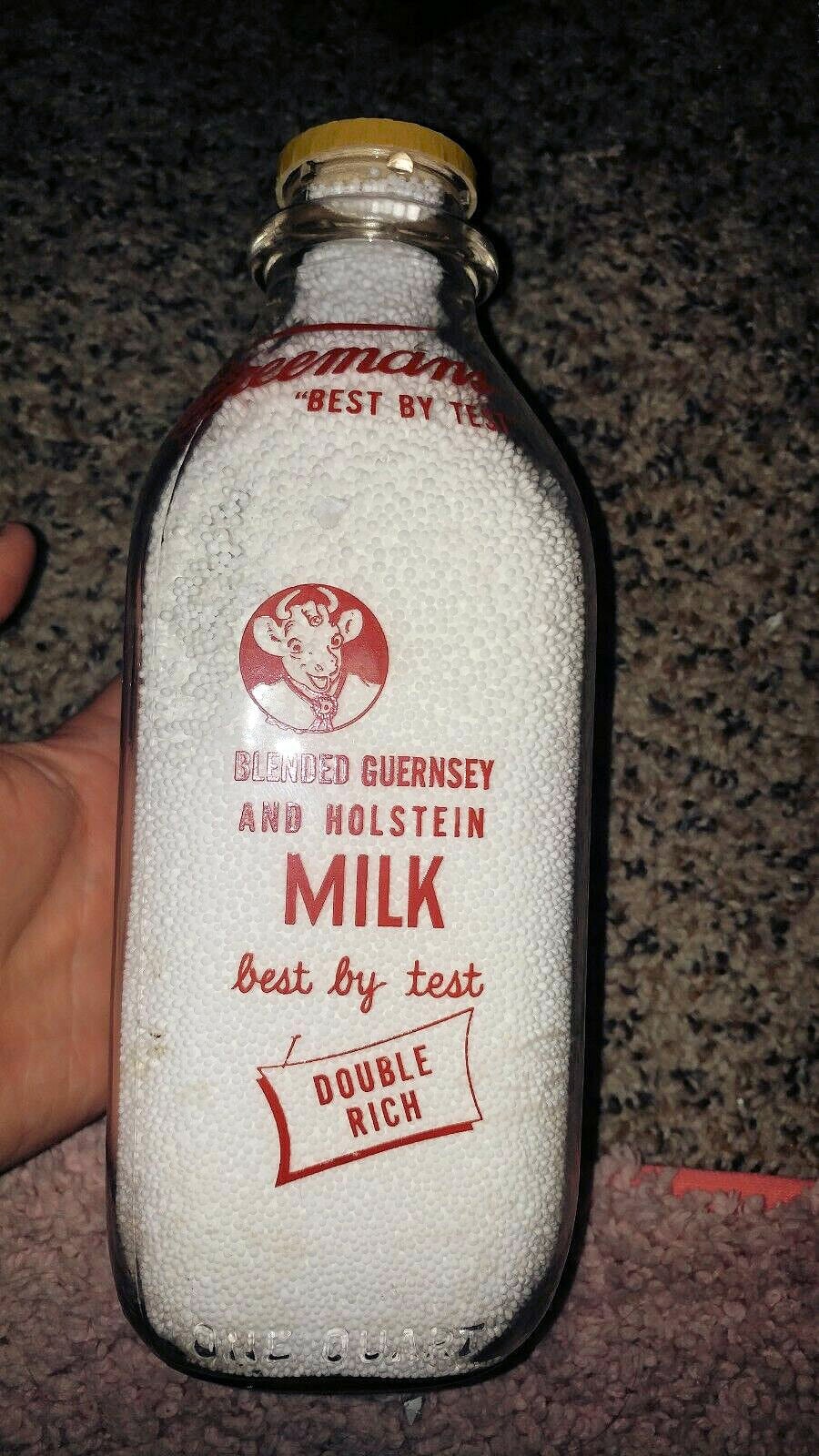 A vintage glass milk bottle labeled “Blended Guernsey and Holstein Milk, Best by Test, Double Rich” sits on a carpet, partially held by a hand. The bottle appears to be filled with a white textured substance.