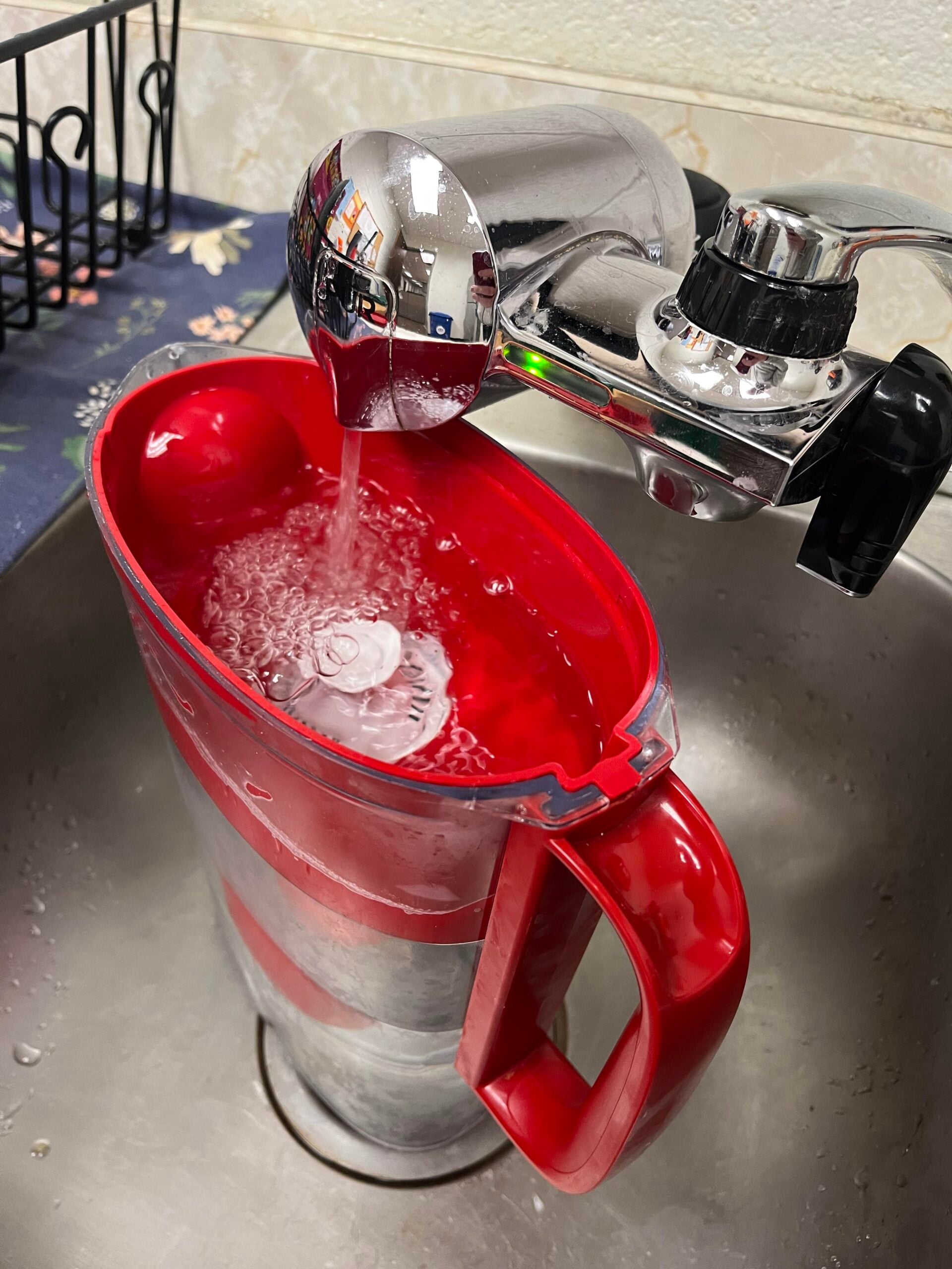 A red plastic pitcher filled with ice cubes is being filled with water from a chrome faucet in a metal sink. The faucet and surroundings are reflected on the shiny faucet surface.