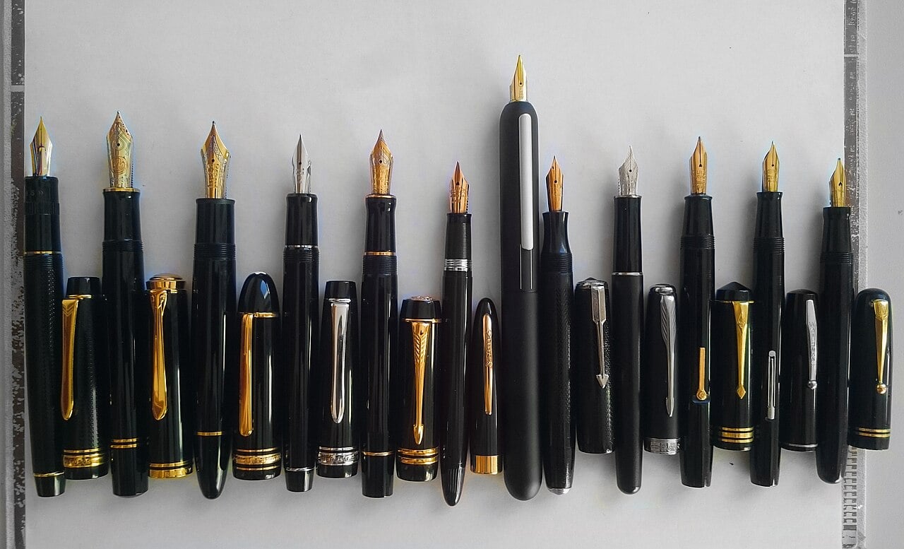 A row of sixteen black fountain pens with gold and silver accents and various nib styles, neatly aligned on a white surface.