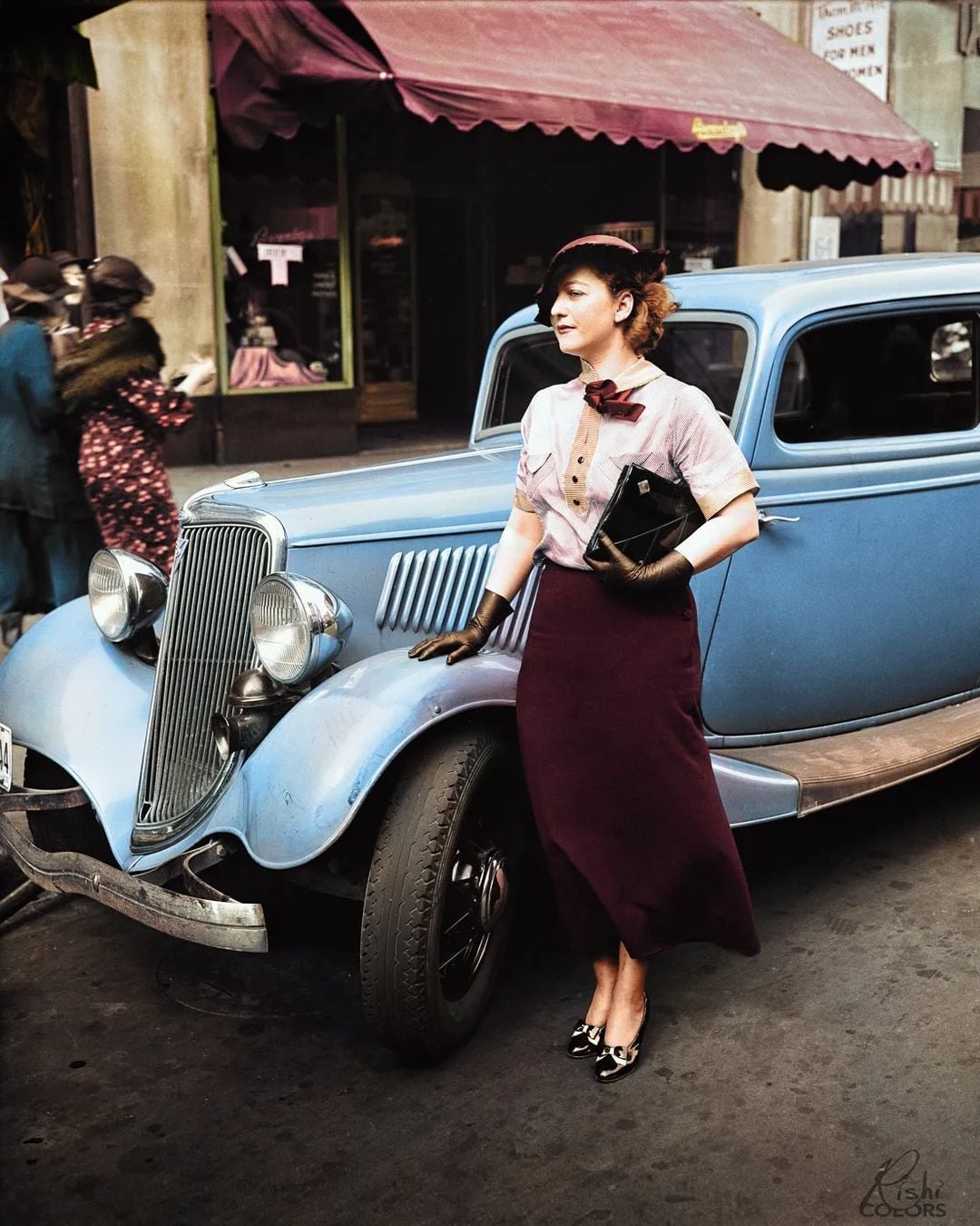 A woman in vintage 1930s attire leans against a blue classic car on a city street, holding a black clutch, with shops and pedestrians visible in the background.