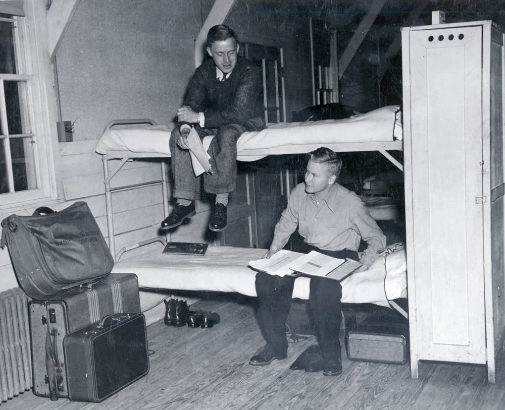 Two young men in a dorm room; one sits on the top bunk, the other on the bottom bunk, both reading papers. Suitcases, shoes, and a locker are visible in the room.