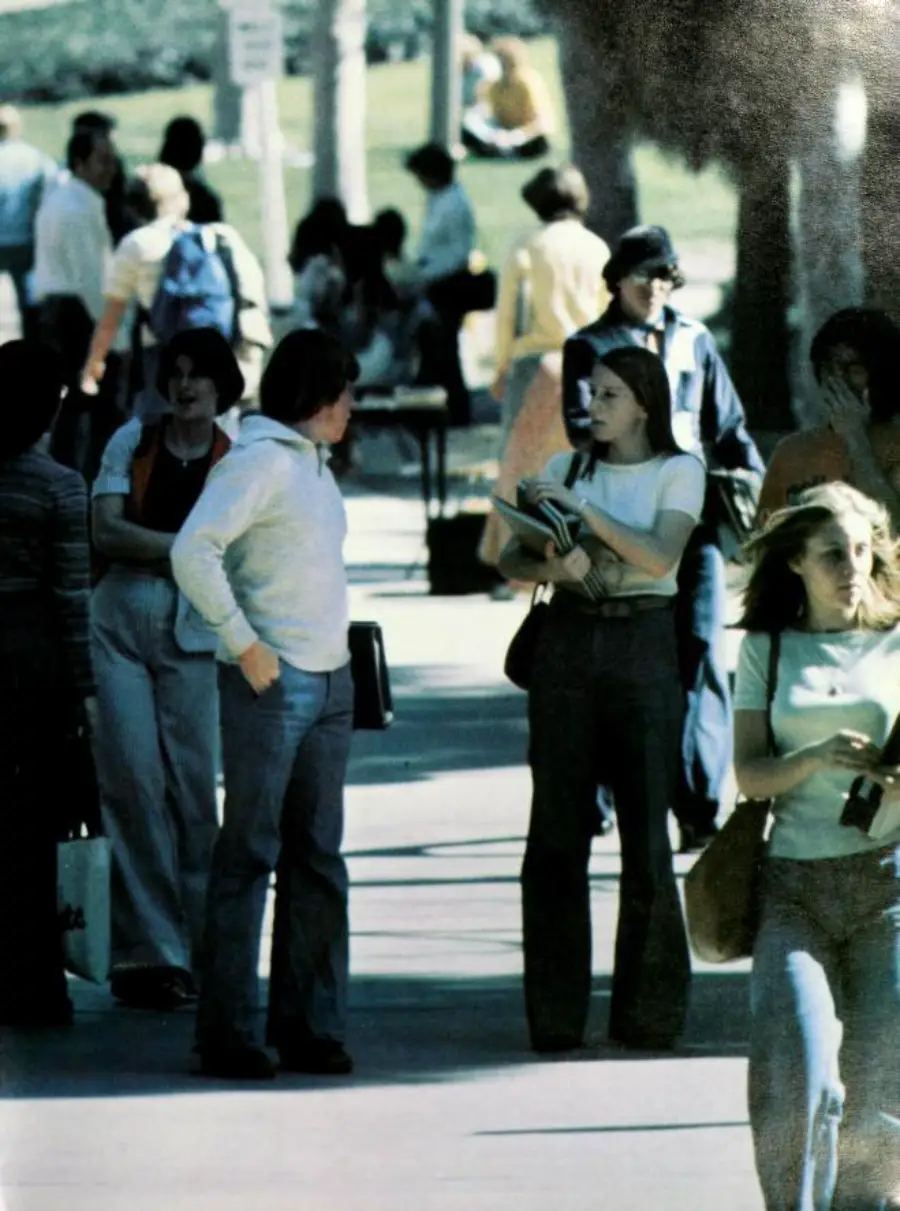 A group of young people walk and talk on a sunlit campus path. Some carry books and bags, and trees provide shade. People are casually dressed, and others sit on the grass in the background.
